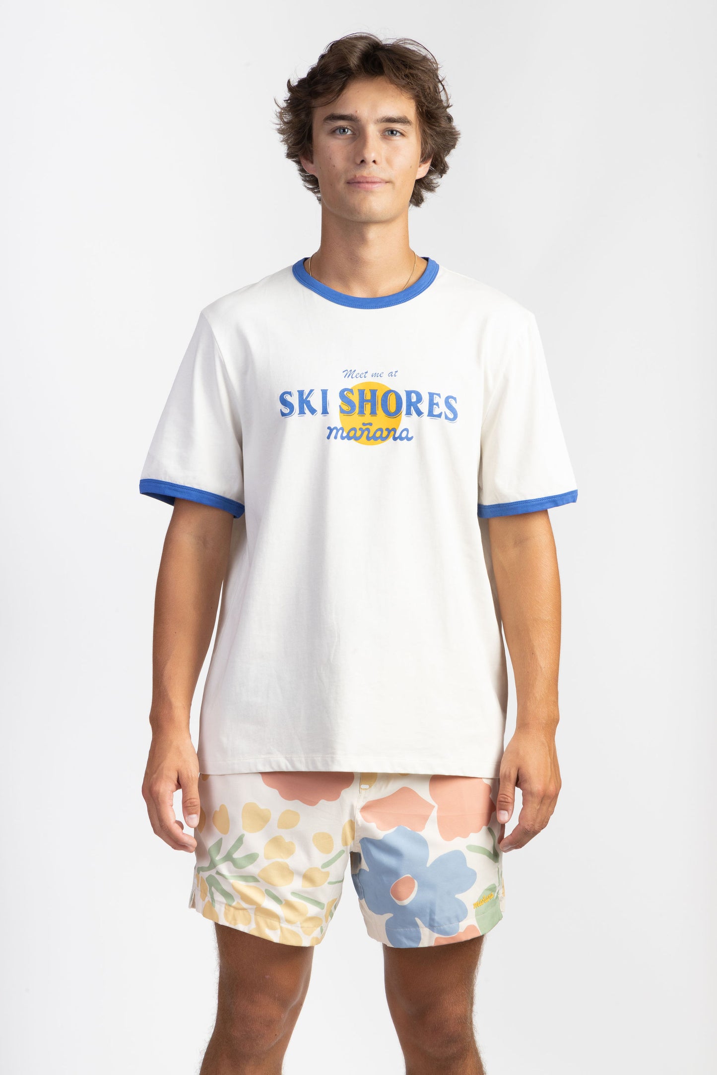 A man wearing a Ski Shores Blue Ringer Shirt having Manana branding with floral shorts