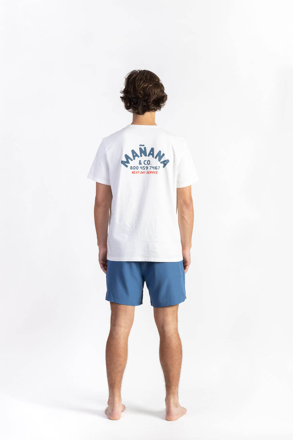 A man wearing a White Shop Tee having Manana branding with blue shorts